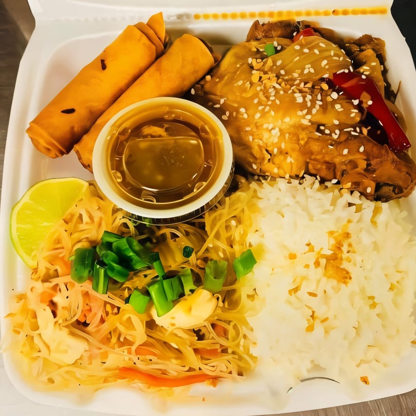 Filipino meal with lumpia, chicken adobo, pancit, rice, and a lime wedge in a takeout container.